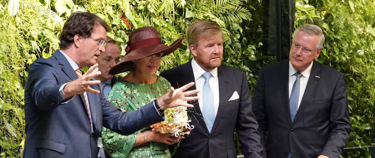 Afbeelding van directeur Jan-Joost Bosman die uitleg geeft aan koningin Maxima en koning Willem met daarnaast commissaris van de koning van Overijssel Andries Heidema met op de achtergrond de groene plantenwand in de fabriek van het bedrijf Auping.